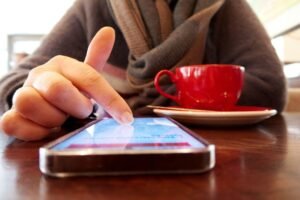 A person uses a smartphone at a wooden table, near a vibrant red coffee cup.