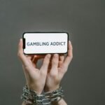 Hands bound in chains hold a phone with "GAMBLING ADDICT" displayed, conveying why internet gambling can be more dangerous.